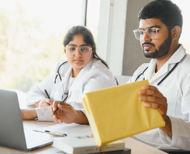 Two medical professionals in white coats, one holding a yellow folder and the other writing, with a laptop and books on the desk.