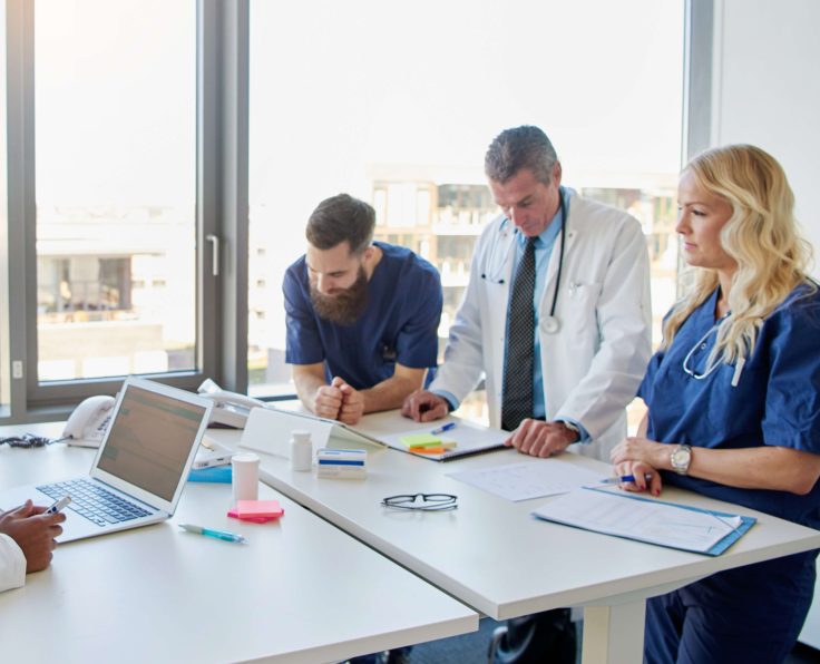 A team of medical professionals in white and blue scrubs reviewing documents and a laptop during a meeting near a window.