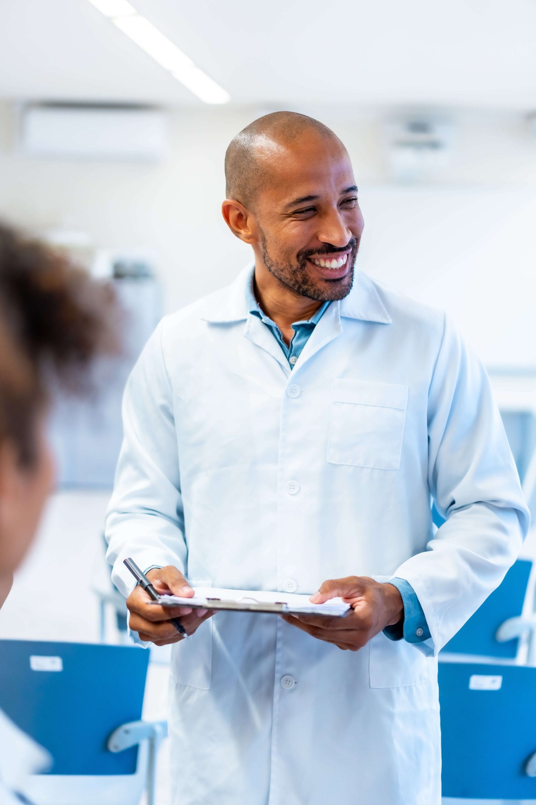 A clinical researcher in a white coat holding a clipboard with a pen, standing in a room with blue chairs and a blurred background.