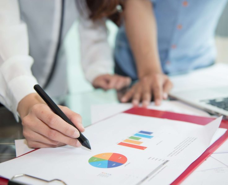 A person writing on a clipboard with a document displaying pie and bar charts, with two others reviewing in the background.