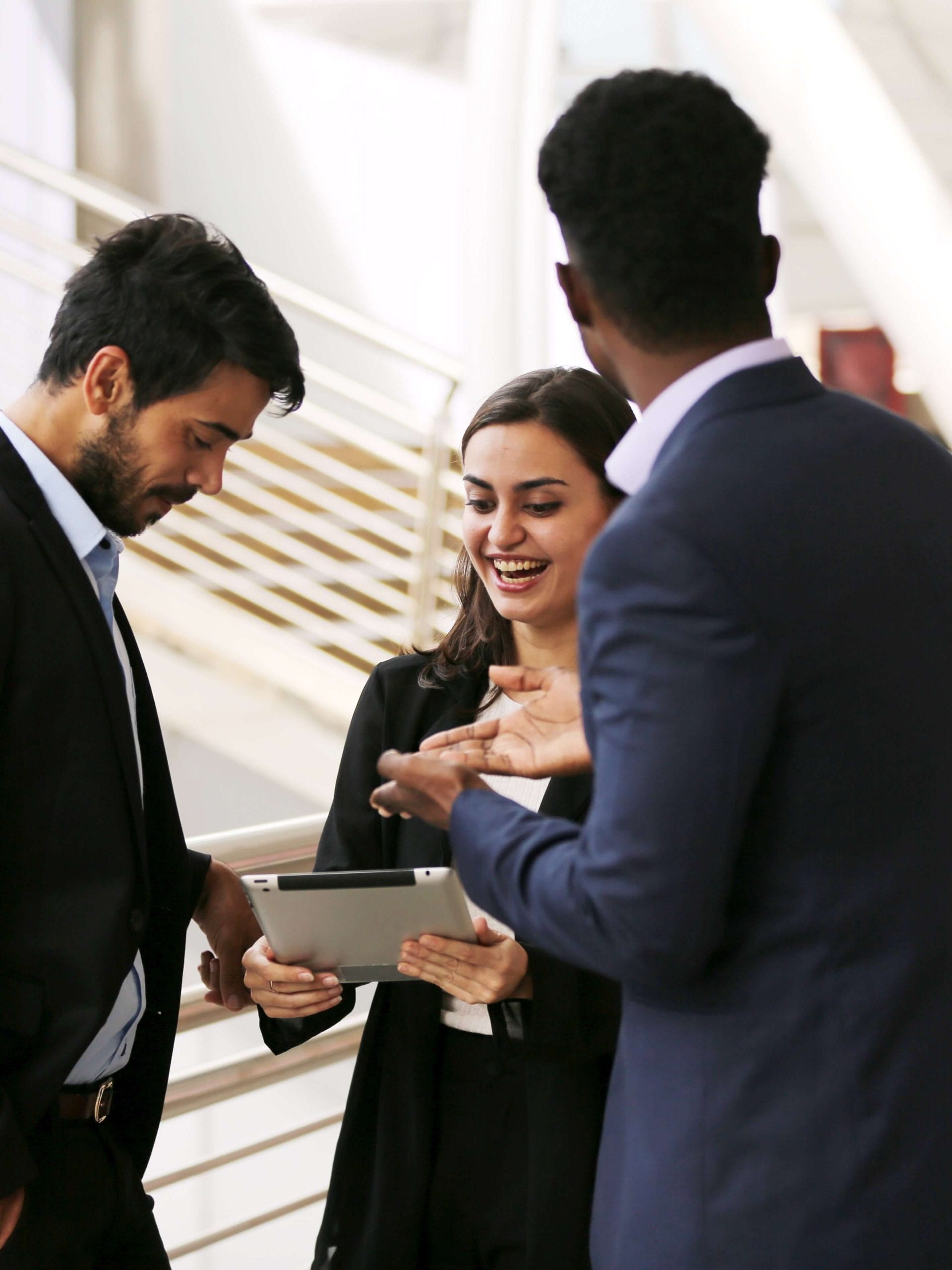 Three professionals in suits engaged in a discussion, with one holding a tablet, standing near a staircase in a modern indoor setting.